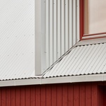 Budova se skládá z lineárního objemu dřevěných panelů, obklopených v přízemí červenými dřevěnými prkny a kovovou trojúhelníkovou střechou z vlnité oceli. Foto: Madara Kuplā