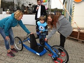 Osmiletý Michal Novotný nasedá na darovaný handbike od Lindabu za asistence své maminky (vpravo), marketingové manažerky Lindabu Petry Ždánské (vlevo) a moderátora akce – herce a režiséra Ondřeje Sokola.