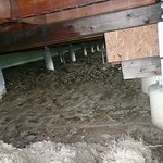 Obr. 6. Crawl space jako bazén na dešťovou vodu, bez možnosti odtoku. Rozbahněné podloží, degradace dřevěných částí vlivem vlhkosti. Nasávání vzduchu z podloží k větrání kanceláří. (Foto: J. Smola).